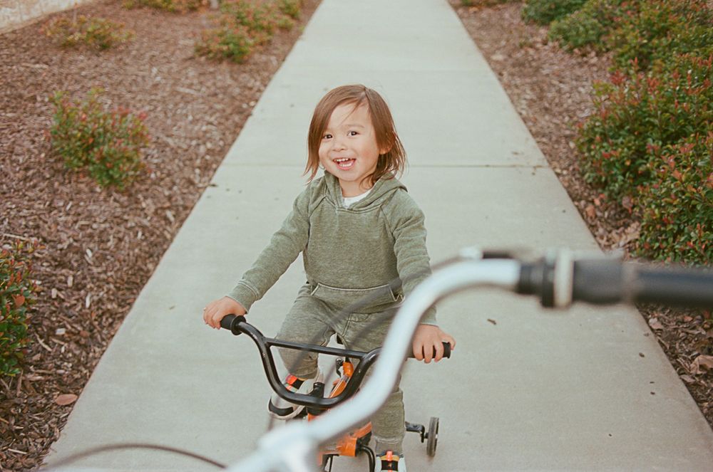 Young child riding a tricycle down a sidewalk pathway during a sunny day.