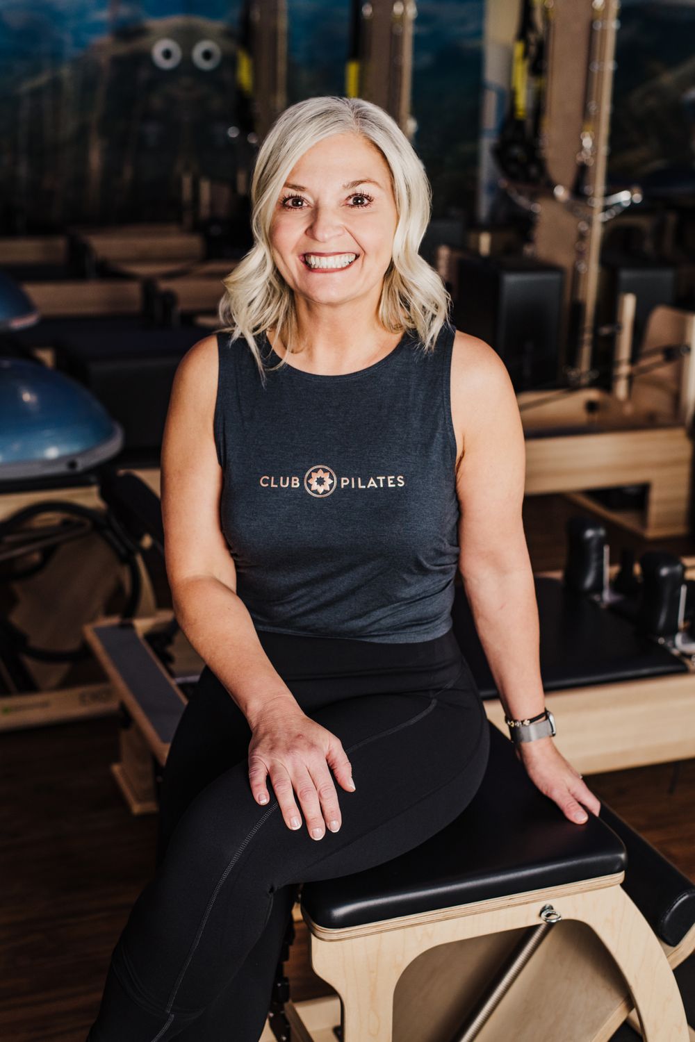 Fitness instructor in Club Pilates tank top sitting on exercise equipment in studio.