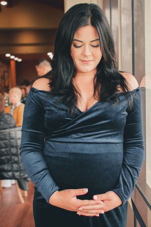 Pregnant woman in navy velvet dress smiles peacefully by window during maternity photoshoot.