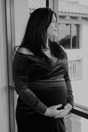 Expectant mother in black velvet dress gazes out window in black and white maternity photo.