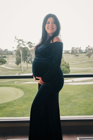 Pregnant woman in black formal dress poses by window overlooking golf course.