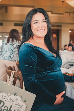 Expectant mother in dark teal velvet dress holding welcome home baby sign at celebration.