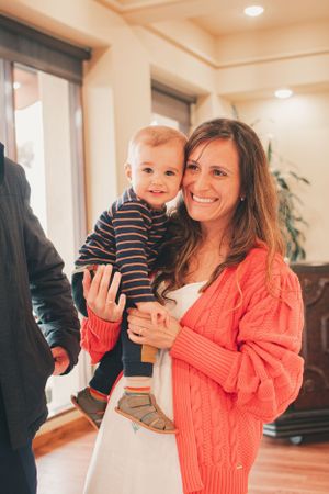 Woman in coral sweater holds small child in striped outfit during indoor celebration.