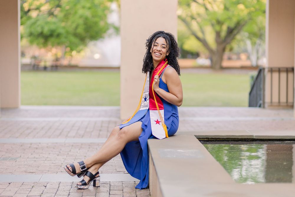 Graduate poses gracefully by campus fountain wearing blue dress and graduation regalia.
