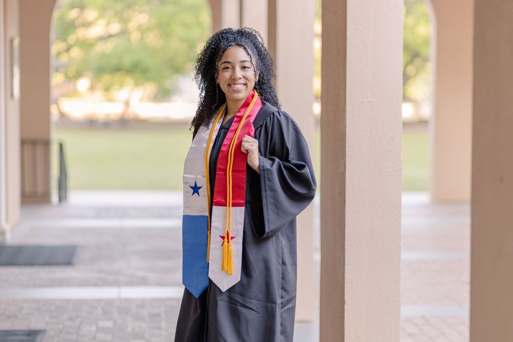 Graduate wearing black graduation gown poses under covered walkway on campus.
