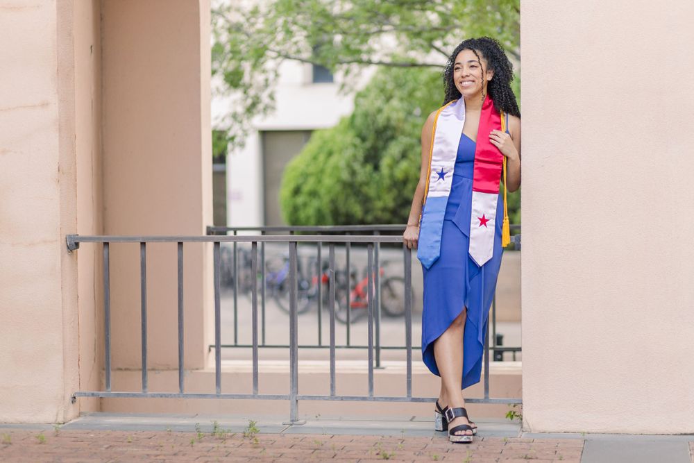A graduate poses in academic regalia against a pink wall with decorative railings.