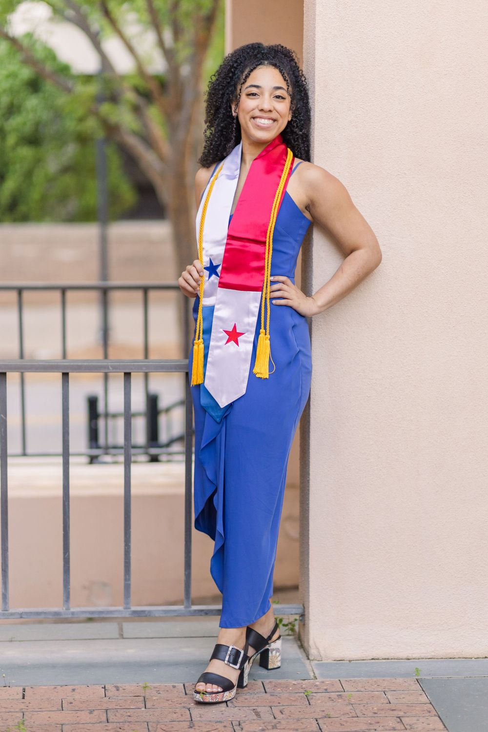 Graduate in blue jumpsuit and graduation stole with red accent poses in front of railing during photoshoot.