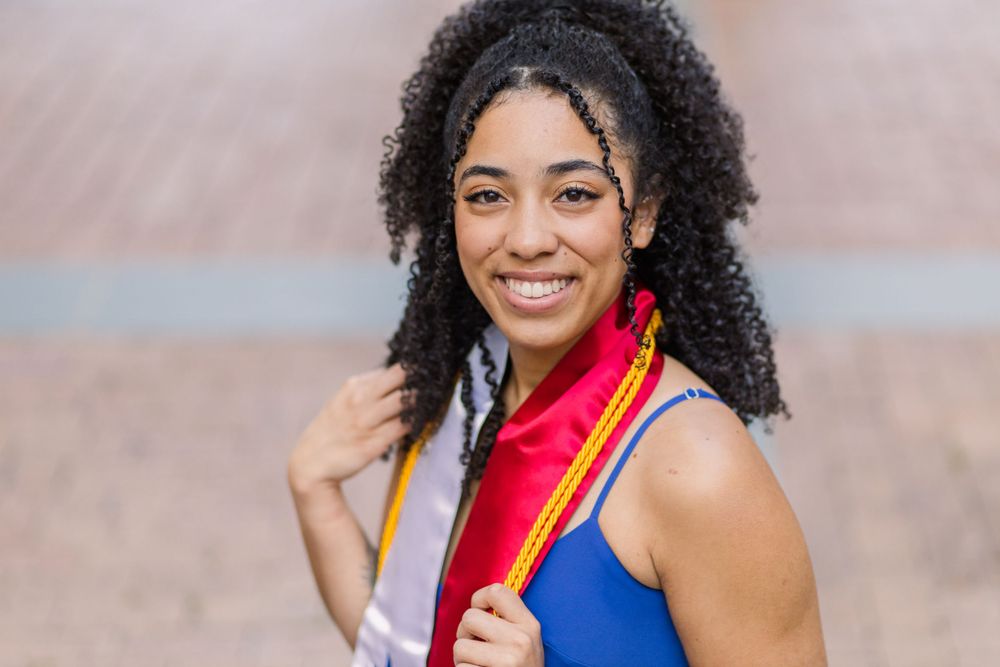 Graduate wearing blue sleeveless dress smiles radiantly while holding graduation stole.