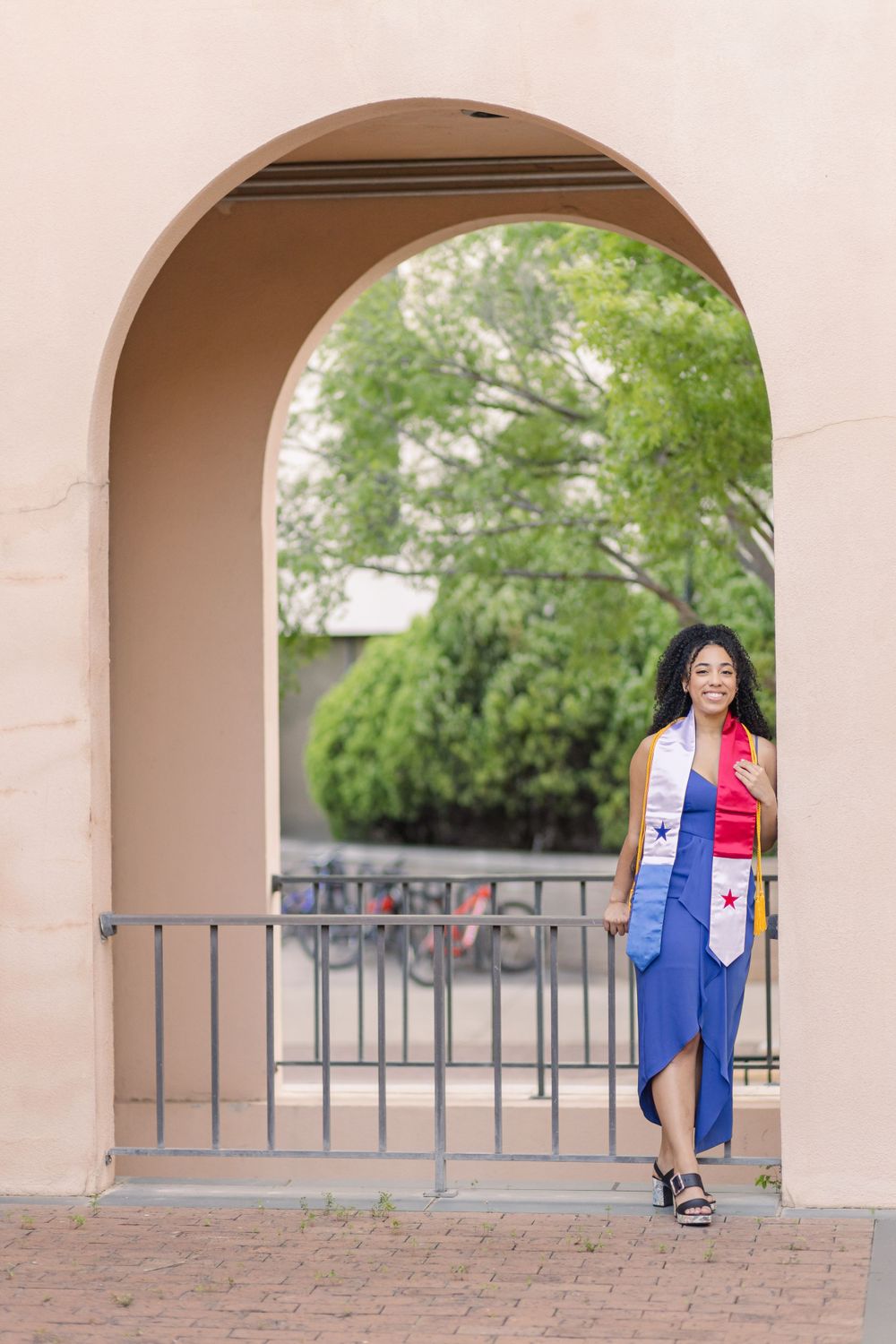 Graduate in blue dress stands under arched doorway during graduation photoshoot.