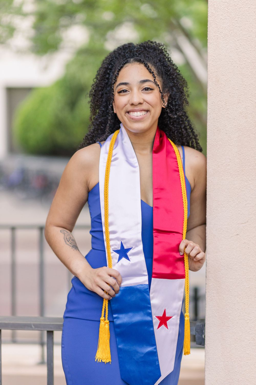 Graduate in blue dress wears decorative graduation stole while posing by campus railing.