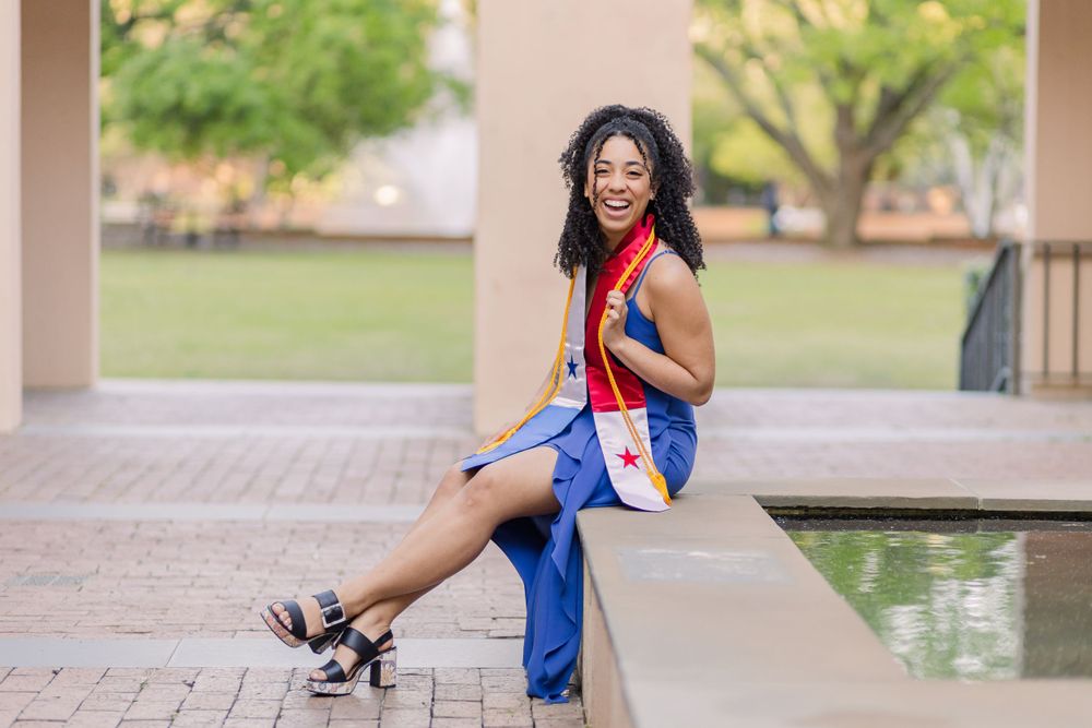 Graduate in blue dress sits elegantly by water feature wearing ceremonial stole.