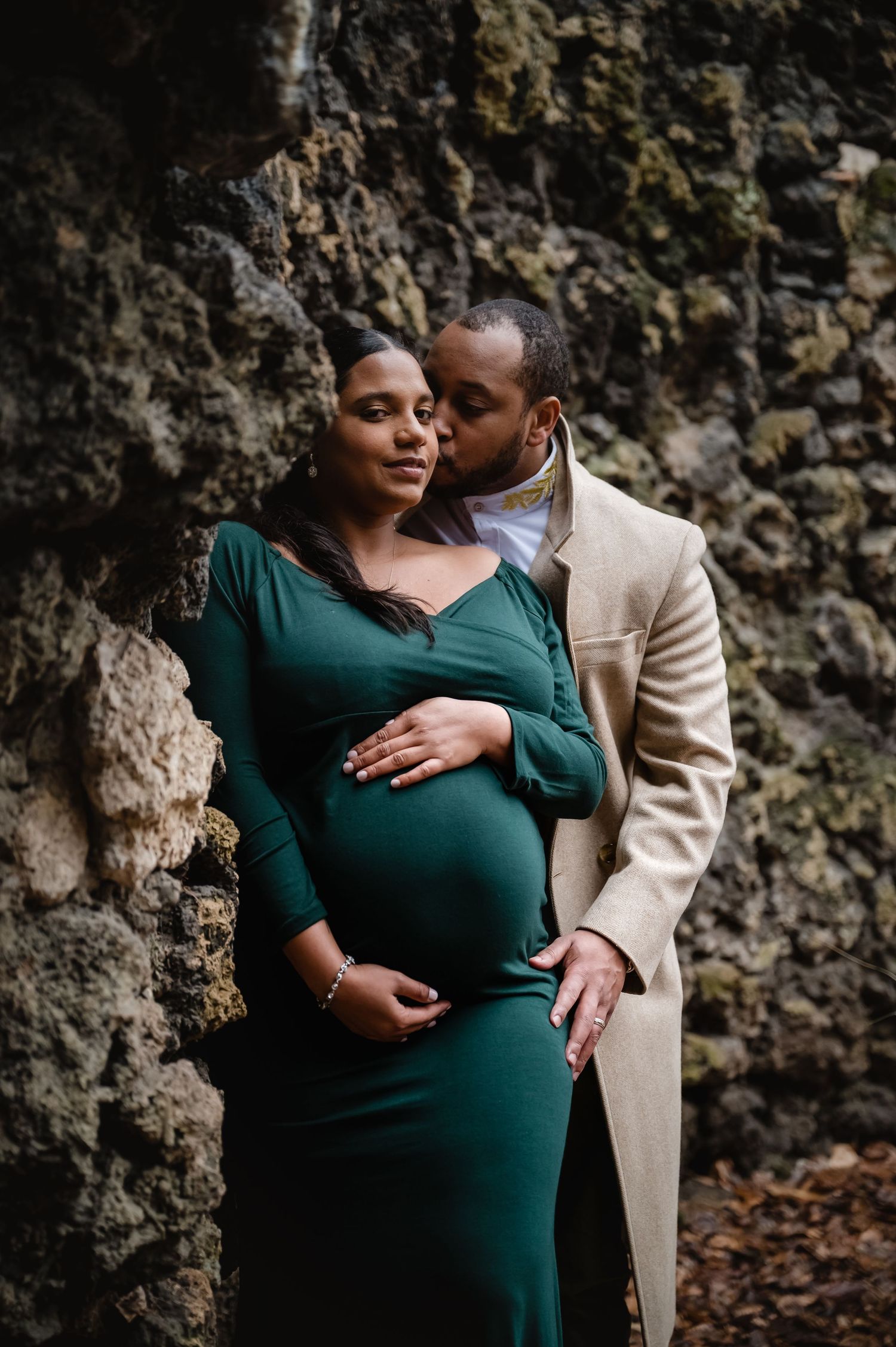 A couple shares a tender moment against a mossy rock wall, woman in a green maternity dress.