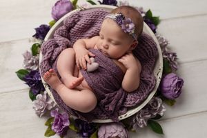 Sleeping newborn in purple wrap surrounded by lavender flowers in round basket.