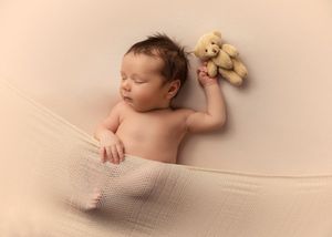 Sleeping newborn posed with small teddy bear on soft beige backdrop.