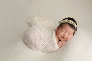 Newborn wrapped in white knit cocoon wearing crystal headband on textured backdrop.