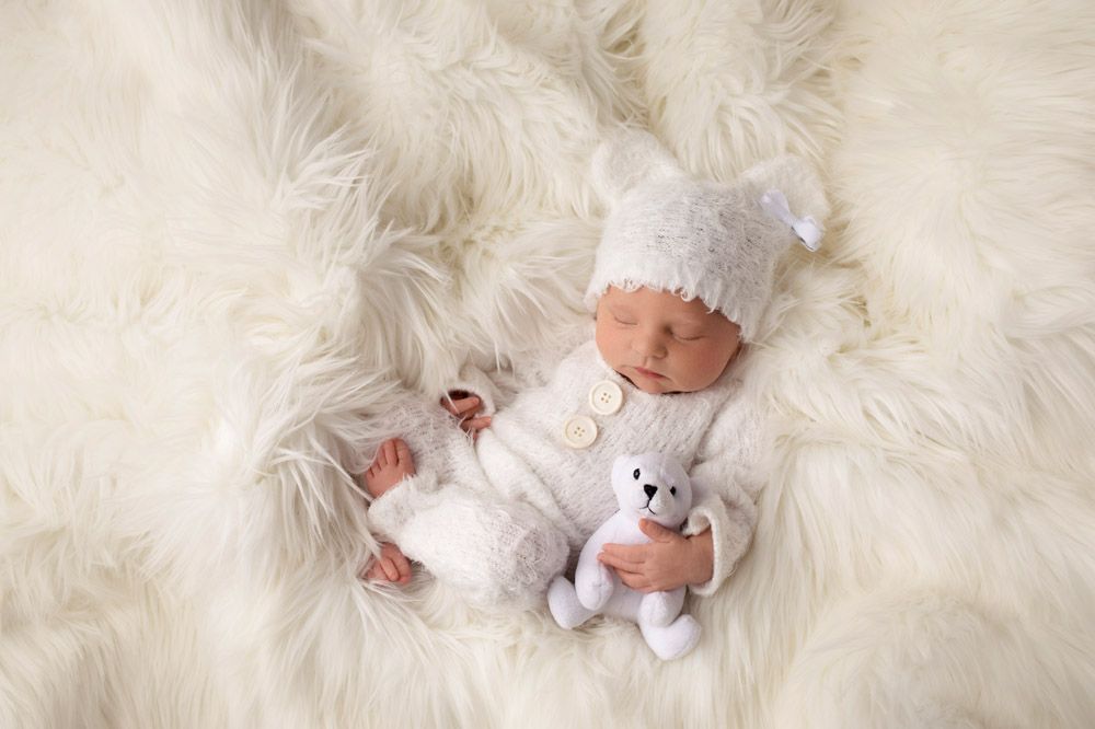 Newborn in white outfit with stuffed animal on fluffy white fur backdrop.