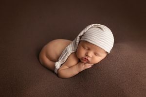 Sleeping newborn wearing white knit hat on brown backdrop in studio portrait.