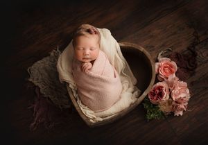 Newborn wrapped in pink swaddle inside wooden bowl with pink roses.