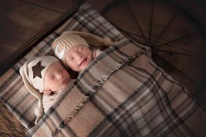 Two sleeping newborns wearing knit hats on plaid blanket.