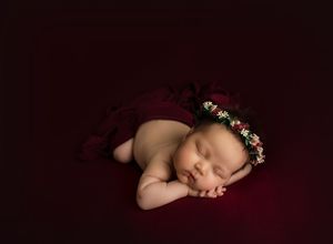 Sleeping newborn with floral crown on dark burgundy backdrop.