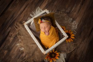Newborn wrapped in yellow swaddle sleeping in white frame with sunflowers.