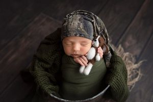 Sleeping newborn wearing camo hat and green wrap while holding small stuffed animal.