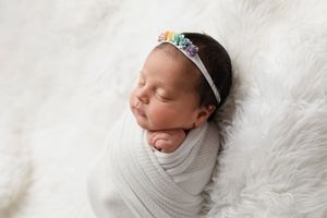 Sleeping newborn wrapped in white cloth wearing delicate floral headband on fluffy white backdrop.