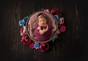 Newborn sleeping in pink basket surrounded by red and blue roses on dark wooden floor.