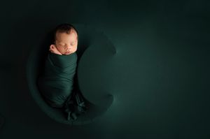Newborn wrapped in dark teal fabric sleeping peacefully on a crescent moon shaped prop.