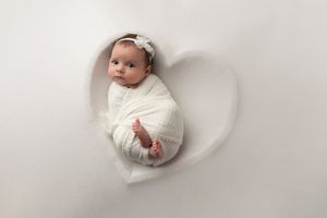 Newborn wrapped in white fabric lying in heart-shaped positioning on white background.