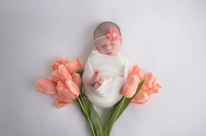 Newborn in white outfit surrounded by coral tulips during spring portrait session.