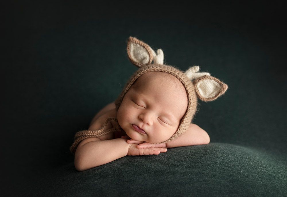 Sleeping newborn wearing knit bunny ears bonnet on dark green background.
