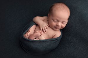 Peaceful sleeping newborn curled up in dark fabric bowl against black background.