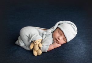 Sleeping newborn in gray outfit with teddy bear on navy backdrop.