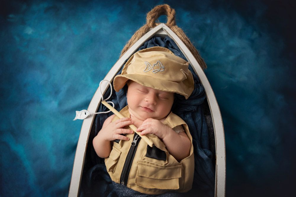 Newborn in sailor outfit sleeping in miniature wooden boat against blue backdrop.