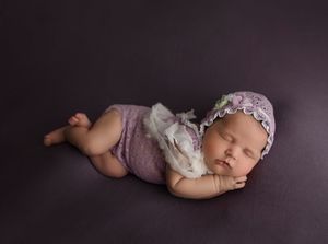 Sleeping newborn in purple outfit with floral headband against dark background.