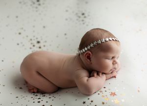 Peaceful newborn wearing sparkly headband resting on white backdrop with gold confetti.