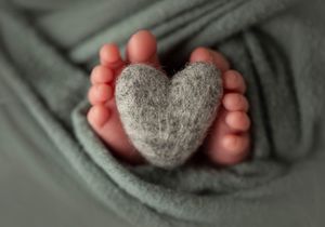 Tiny baby feet holding gray heart-shaped plush toy.