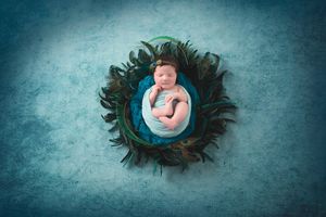 Newborn in blue wrap nestled in feathered nest on textured teal backdrop.