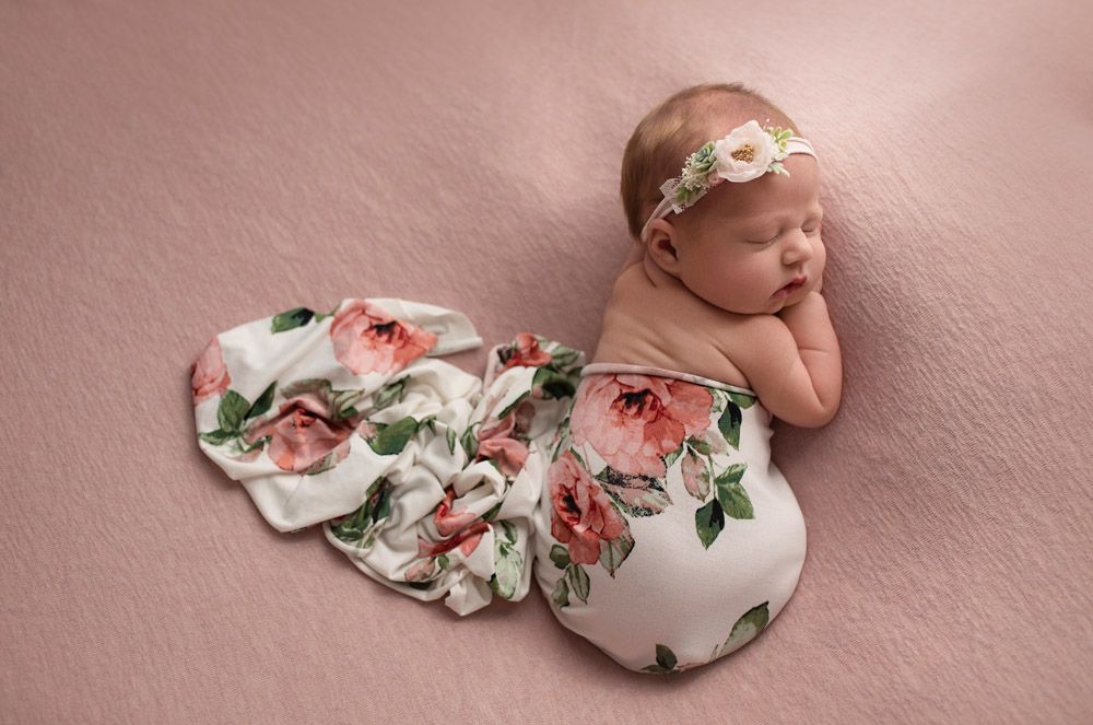 Sleeping newborn in floral dress and headband on pink background.