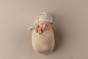 Newborn baby wrapped in beige swaddle wearing striped knit cap during professional photography session.
