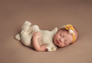 Sleeping newborn in cream outfit with yellow floral headband on brown background.