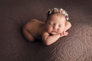 Peaceful newborn with floral crown resting on textured brown fabric during photo session.