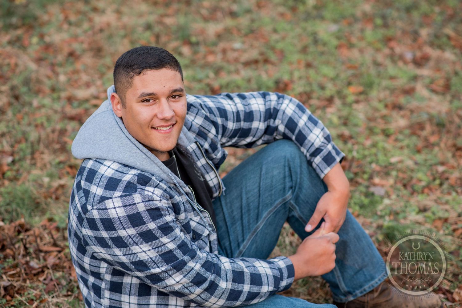 A person in plaid sitting on autumn grass smiles warmly at the camera.