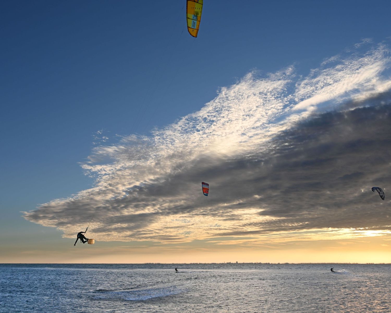 Kitesurfers soar against a dramatic sunset sky over ocean waves.