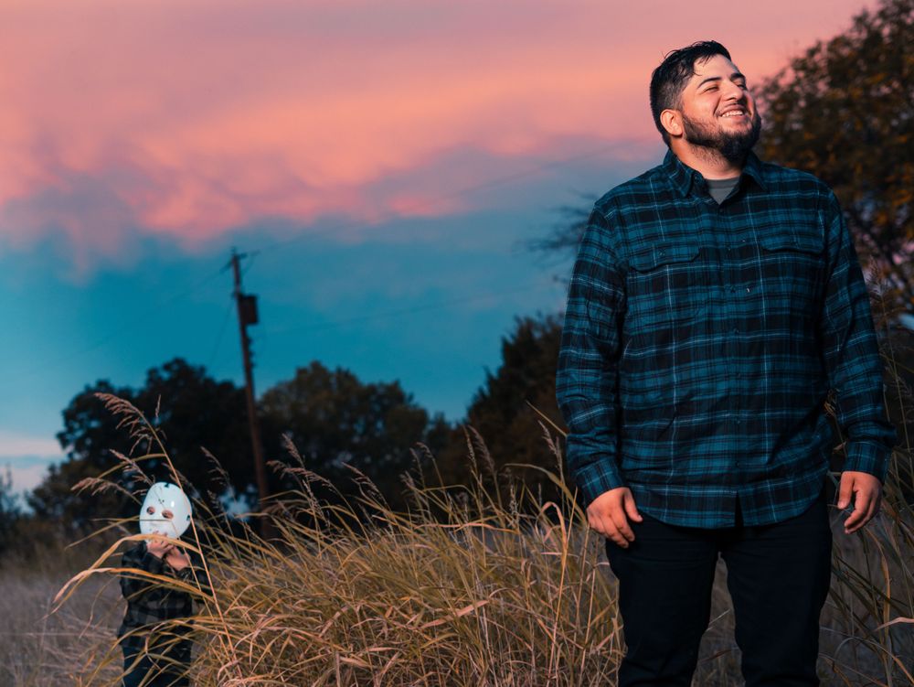 Person in plaid flannel shirt stands in tall grass against vibrant sunset sky.