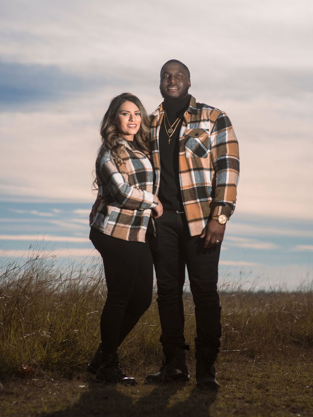 Couple wearing matching plaid jackets pose together in field at sunset.