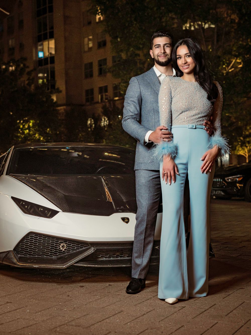 A stylish couple poses in formal attire next to a white Lamborghini sports car at night in an urban setting.