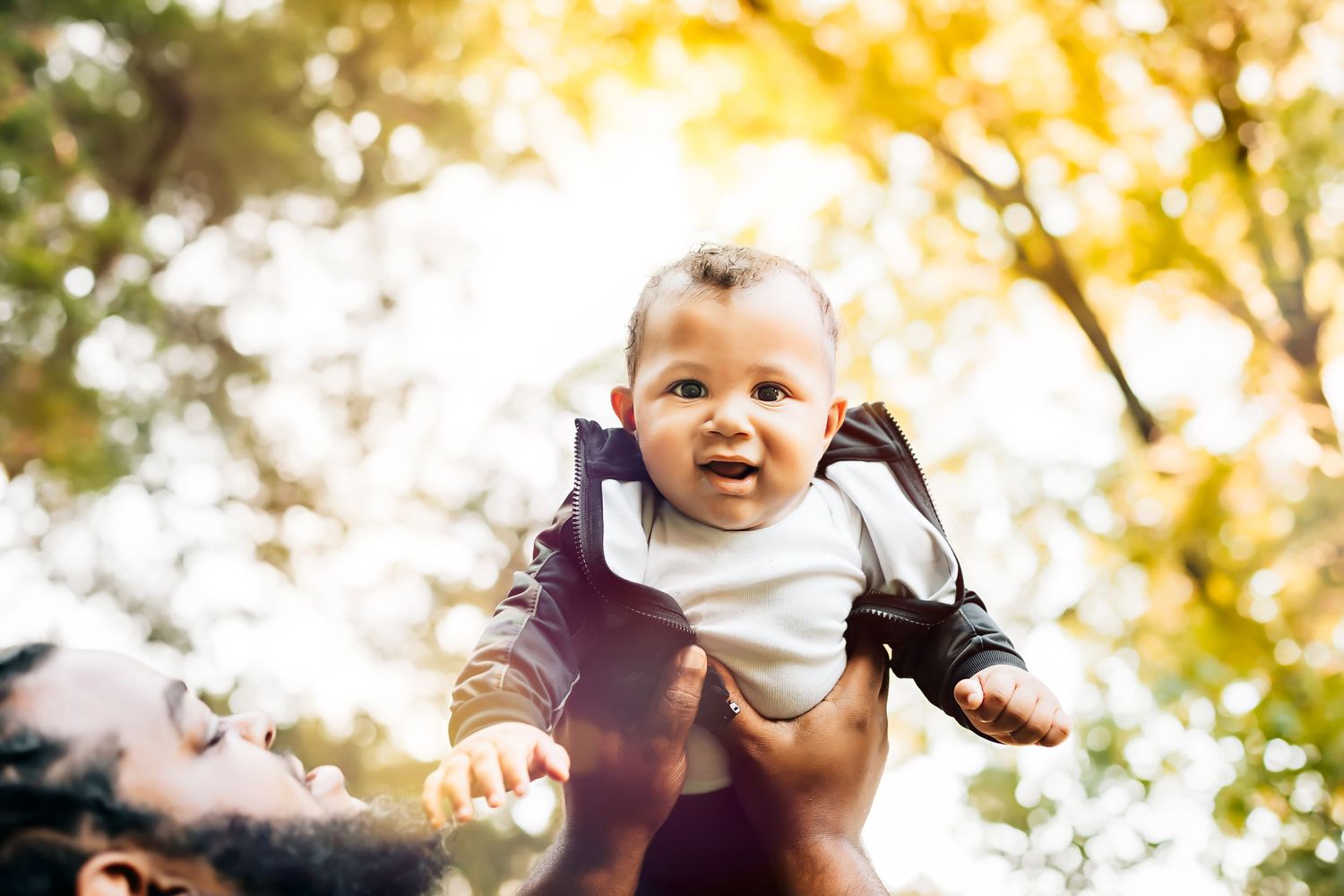 A joyful baby wearing black jacket is lifted high in golden sunlight.
