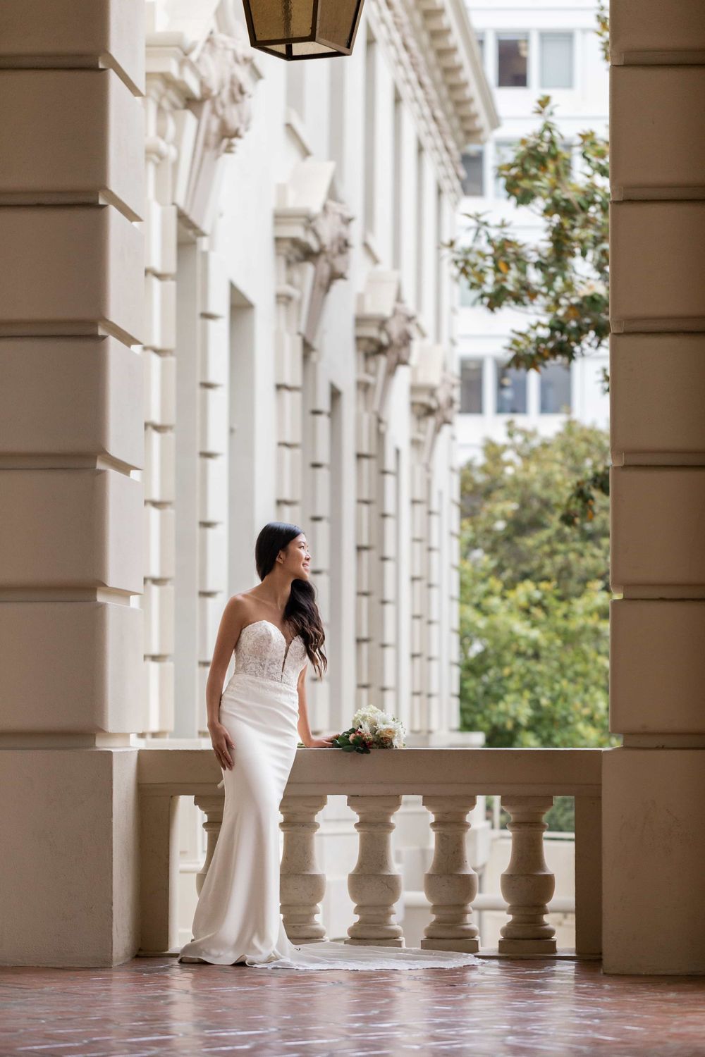 Bride in strapless lace and crepe wedding gown holds bouquet on elegant columned balcony.